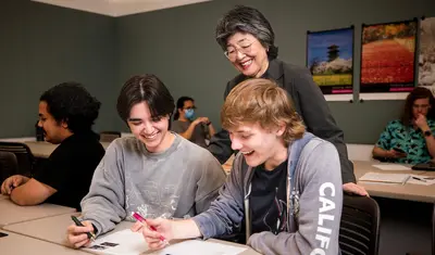 Two students working together on a worksheet with the professor standing behind them supporting them and smiling.