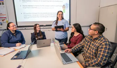 A group of students sit around a table with laptops while a student stands at the front of the classroom holding a tablet and leading a discussion, with a projected slide on cognitive psychology displayed behind her.