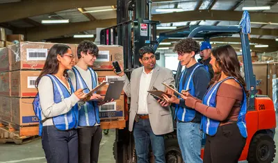 Four students looking at their clipboards in a warehouse with the professor standing next to them advising them.