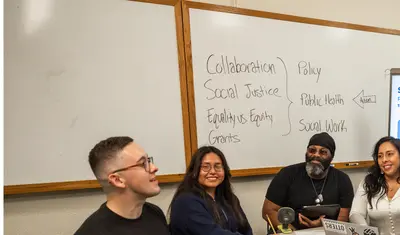 Group of students listening and looking at one student speak at a table in a classroom.