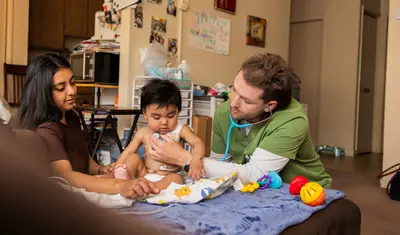 A healthcare student provides care to a child while engaging with a family in a supportive home setting.