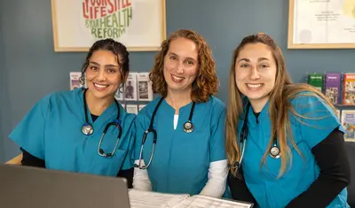 A group of three nursing students in scrubs with stethoscopes around their necks sitting down and smiling at the camera