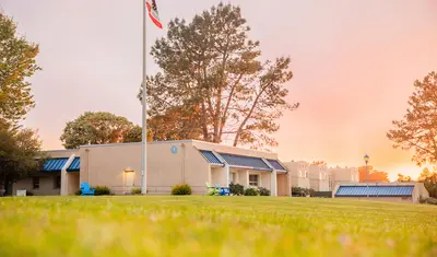 A campus lawn glows at sunset with buildings, trees, and U.S. and a California flag under a sky filled with golden clouds.