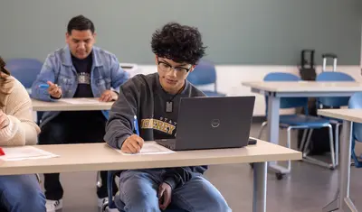 A student works independently in a classroom, using a laptop while completing an assignment.