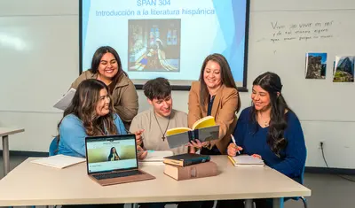 Instructor and students gathered around a table reading and discussing a book during a Spanish literature class with a presentation screen behind them.