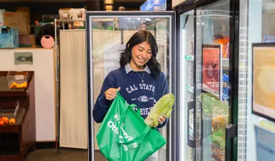 Student smiling while holding fresh produce and an Otter Care bag inside the pantry.