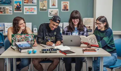 A group of four students sitting at a table collaborating with computers, books, and papers on the table.