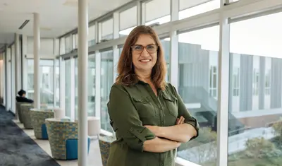 A ���ӽ紫ý Humanities and Communication faculty member stands with arms crossed in a bright campus hallway, smiling in front of large windows and modern seating areas.
