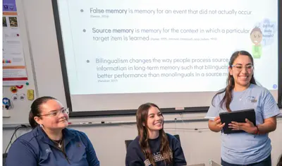 A group of students sit around a table with laptops while a student stands at the front of the classroom holding a tablet and leading a discussion, with a projected slide on cognitive psychology displayed behind her.