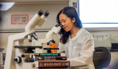 A student wearing a lab coat sitting down looking through a microscope
