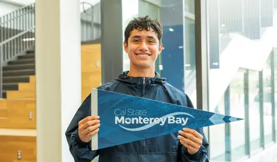Student smiling proudly holding a CSUMB pennant inside a CSUMB logo.