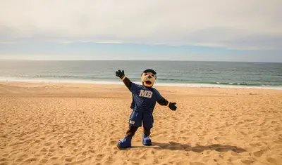 The campus mascot stands on a sandy beach with the ocean behind, highlighting the university’s coastal setting and school spirit.