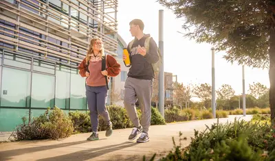 Two ���ӽ紫ý college students walk and chat on a sunny campus path, carrying backpacks and a water bottle near a modern building.