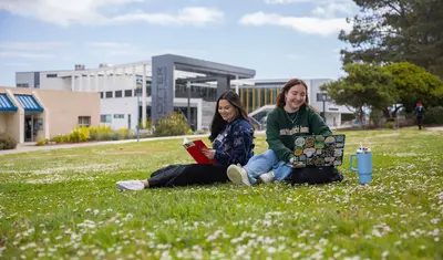 Two CSUMB students sitting on the grass in Main Quad with the Otter Student Union (OSU) in the background