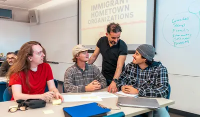 Three students sitting at a table in a classroom with the professor advising them on their work.