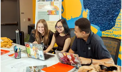 Three students sitting at a table smiling and looking at a computer together