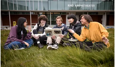 Multiple students sitting on the grass all reading from the same book and smiling.
