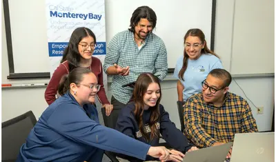Three students sitting at a table looking at the a computer in the middle of them, with three students standing behind them also looking at the same computer.