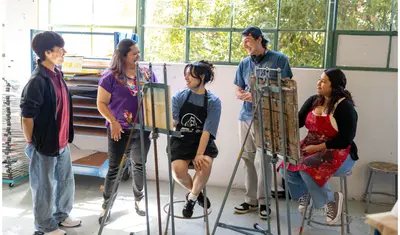 Three students standing and smiling around two students who are sitting and painting on canvases.