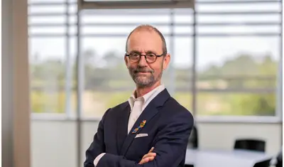 Professor in a suit poses confidently with arms crossed in a bright classroom.