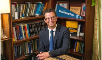 Professor sitting in front of a book shelf, looking up at the camera smiling with his hands on the table in front of him.