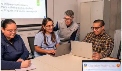 Three professors sitting at a table on their computer and taking notes while discussing with a student also sitting at the table