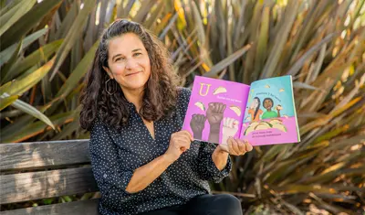 A woman sitting on a bench outdoors smiles while holding open a colorful children’s book featuring illustrations of raised fists and people eating tacos, with text in both English and Spanish.