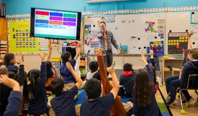A teacher stands at the front of an elementary school classroom, pointing toward students who are seated on the floor with their hands raised.