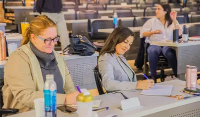 Students at California State University, Monterey Bay (CSUMB) take notes and engage during a classroom or workshop session in a modern lecture hall. The atmosphere is collaborative and professional, emphasizing active learning and participation.