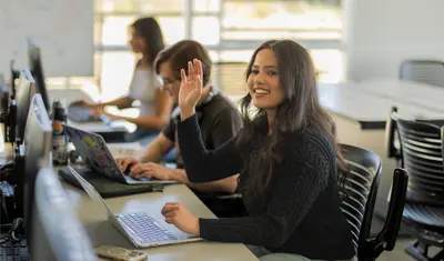A group of students working while one has their hand raised asking a question