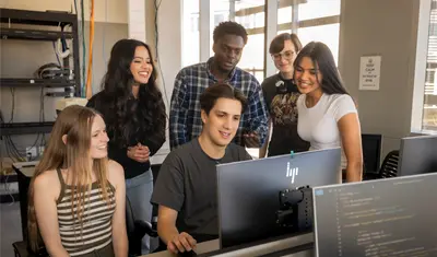 A group of students looking at a students desktop while they work on it