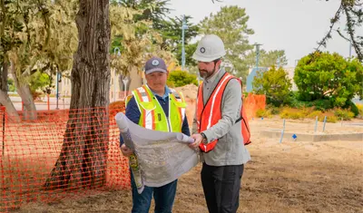 wo CSUMB facilities professionals review blueprints at an outdoor construction site, standing under trees near orange safety fencing and discussing the project layout.