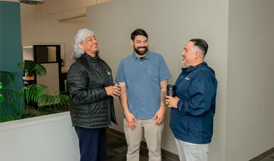 Three CSUMB finance department colleagues stand together in an office hallway, laughing and holding coffee mugs during a casual morning conversation.