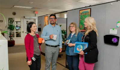 Four CSUMB HR staff members enjoy a casual coffee break together in a collaborative office space, laughing and chatting with colorful mugs and team materials in hand.
