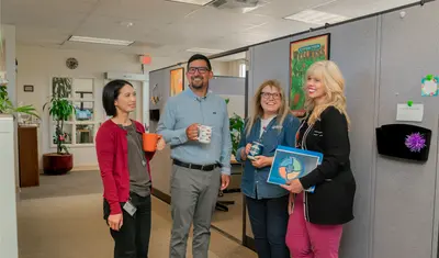 HR colleagues at CSUMB continue a cheerful conversation in the hallway, holding coffee mugs and standing next to personalized cubicle walls decorated with wellness posters.