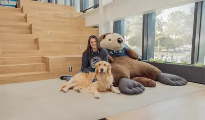 Blue the therapy laying down in front of the large Monte plushe in the OSU with his owner sitting behind him.