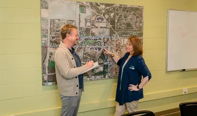 A staff member points at a large wall-mounted campus map while another takes notes. They appear to be discussing safety logistics.