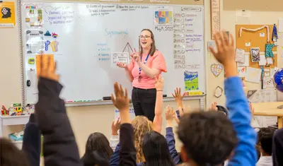 Teacher stands at the whiteboard while students raise their hands in class