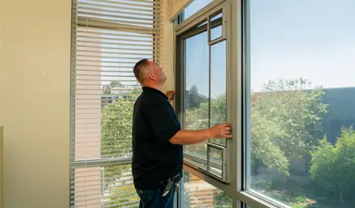 A technician checks a window latch and frame inside a residence hall, inspecting for safety and operability.