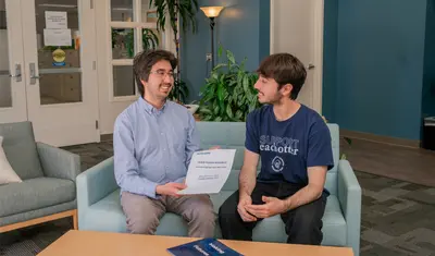 An HR employee and a ���ӽ紫ý student sit on a couch in the HR reception area reviewing a new hire packet for student assistants, surrounded by welcoming decor and indoor plants.