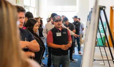 Student researcher presents her agricultural science project at CSUMB’s AGPS Presentation. Audience members stand close, engaging with the poster content.