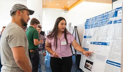 A student at CSUMB explains a research poster on agricultural science to a group of attendees during the AGPS Summer Presentation. The student gestures toward the poster while others listen closely.