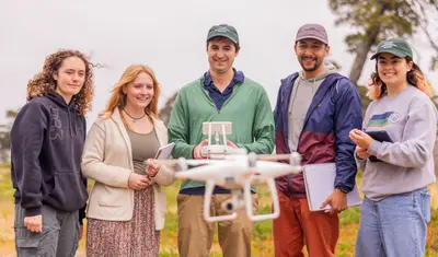 A group of four AES students and a professor standing in the grass while the professor is hovering a drone in front of them