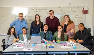 A group of students posing for a picture together in front of a desk, half of them sitting and half of them standing behind them.
