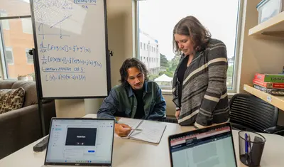 A student explaining a question to a professor who is standing next to him helping him on a paper.