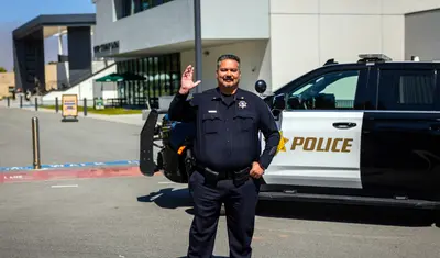 A male ������app UPD  officer smiles and waves in front of the police SUV. Campus buildings and a clear sky complete the friendly scene.