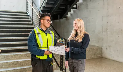 A community service officer shares a flyer and campus map with a smiling student in a stairwell area of a modern campus building.