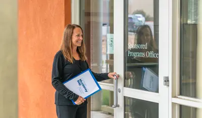 A staff member opens the door to the Sponsored Programs Office while holding a CSUMB Sponsored Programs folder and smiling.