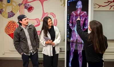 A student is giving a presentation using a digital anatomy board to two other students