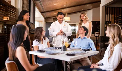A chef in a white coat stands beside a restaurant table speaking with a group of students seated around a formally set dining table in an upscale dining space.